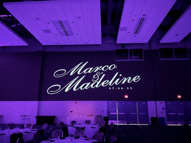 A monogram projector casting a couple’s names and wedding date in large script lettering across a ballroom wall lit in purple