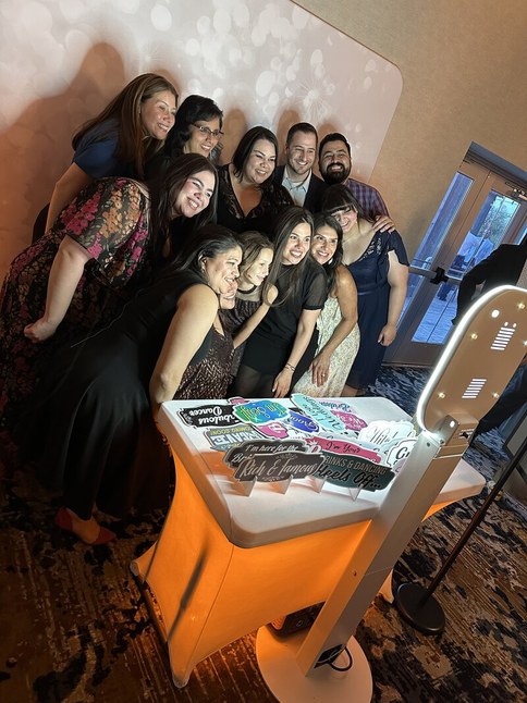Guests crowded together laughing in front of the Signature Photo Booth, beside a table laid with printed photo strips and props